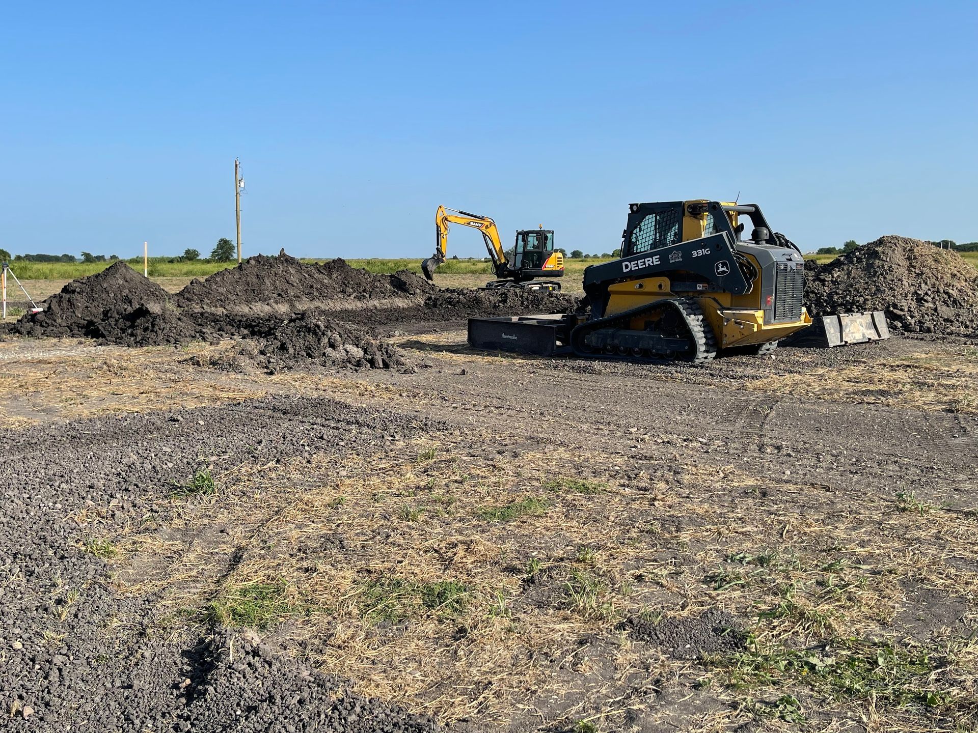 A yellow skid steer and an excavator operate on a dirt construction site under a clear blue sky.