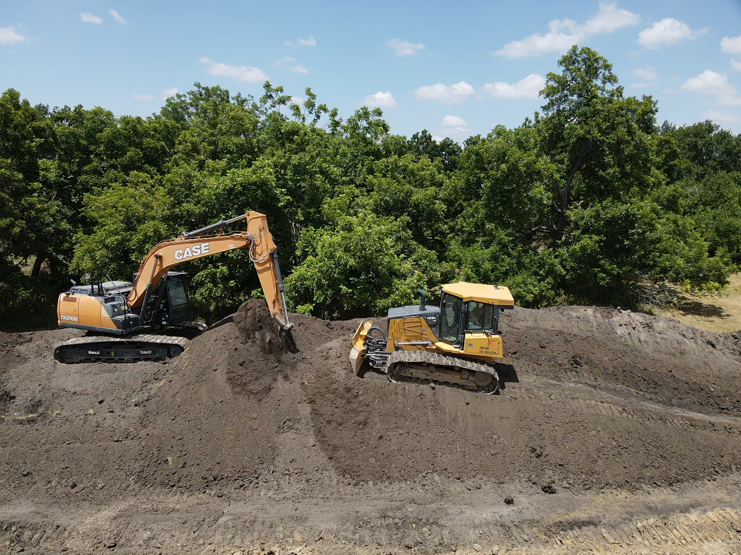 A yellow excavator and a bulldozer work on a large pile of dark soil in a field bordered by trees.