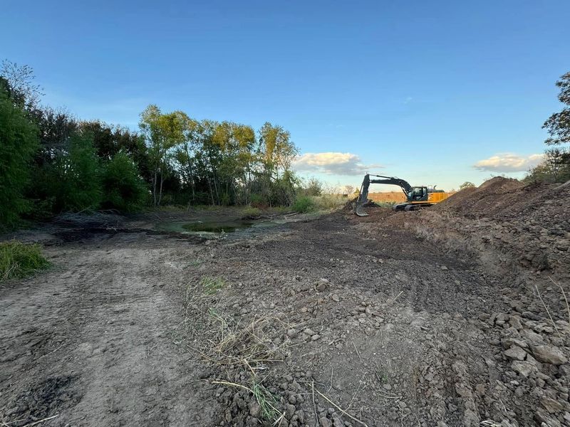An excavator sits on a dirt mound in a cleared, rural area with trees under a blue sky.