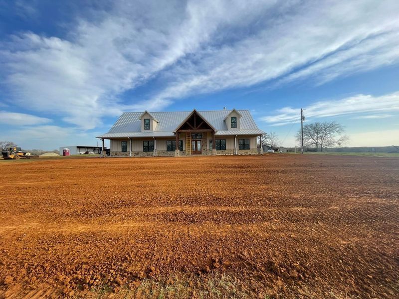 A single-story home with a metal roof and a large covered porch, situated behind a expansive, freshly graded red dirt yard.