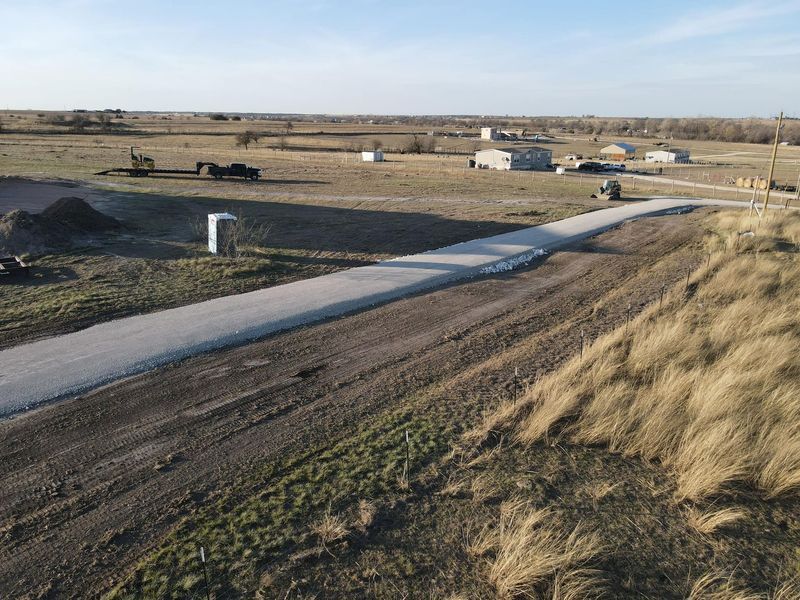 A paved path winds through an open, dry, grassy field with several buildings and vehicles in the distance.
