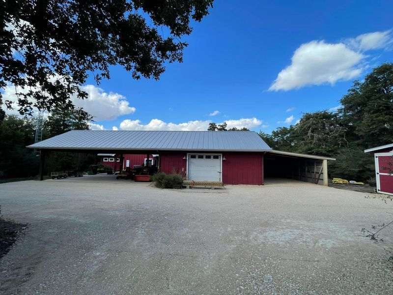 A red barn with a gray metal roof, a white garage door, and a gravel driveway under a sunny blue sky.