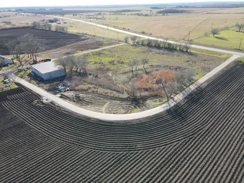 An aerial view of a rural corner lot with a metal building, trees, and a small pond, bordered by plowed dark soil fields.
