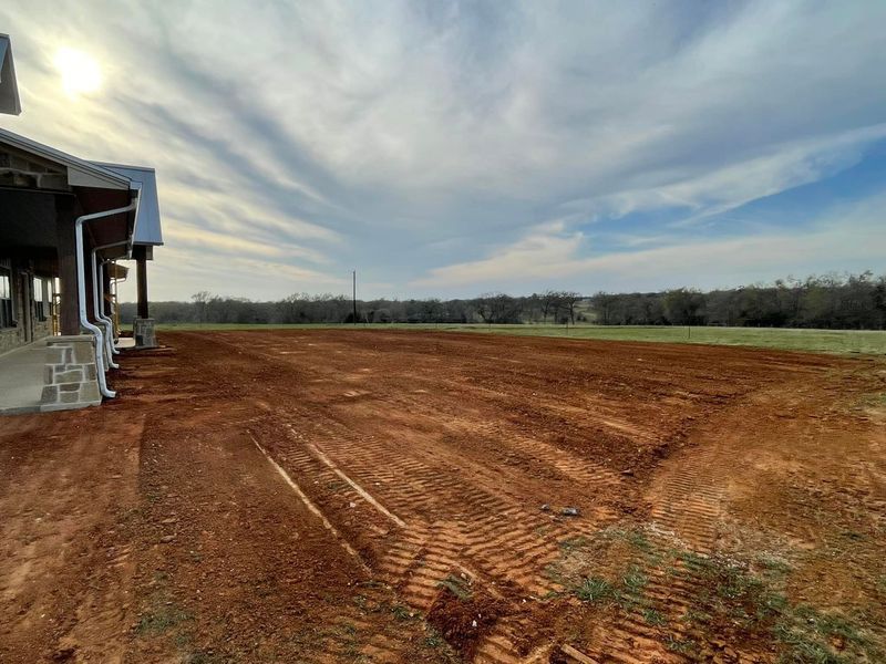 Newly graded red dirt stretches from the side of a house toward a grassy field under a cloudy, late afternoon sky.