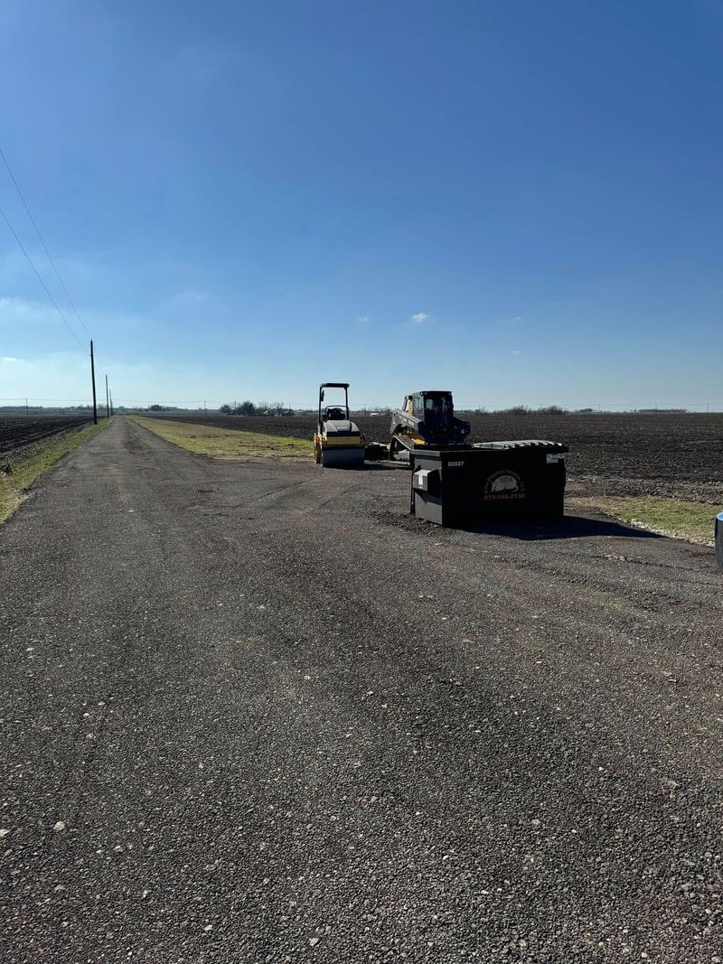 Construction equipment, including a small roller and a tractor, parked on a gravel path next to a plowed field.