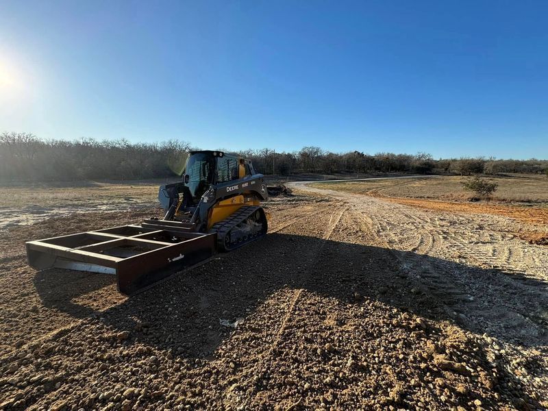 A yellow skid-steer loader with a large metal frame attachment sits on a dirt construction site under a clear blue sky.