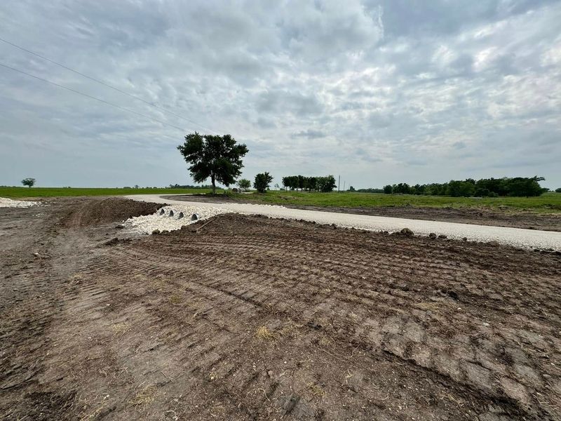 A light-colored dirt path winds through a construction site, leading toward a solitary tree under a cloudy sky.