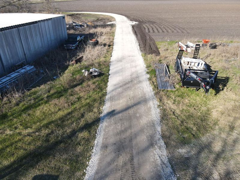 An aerial view of a long, light-colored gravel driveway running between a metal shed and a field, with a trailer parked.