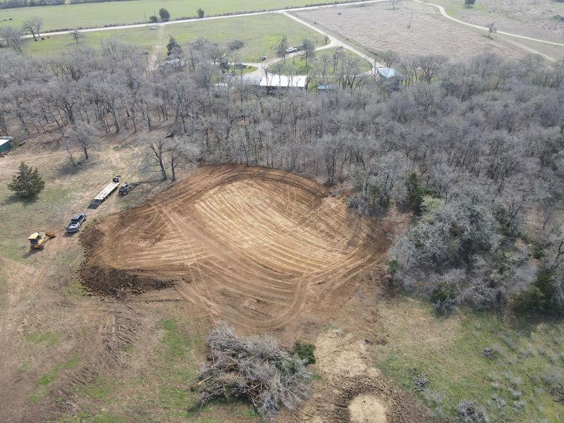 An aerial view of a large, excavated dirt pond area in a rural, wooded landscape with construction equipment nearby.