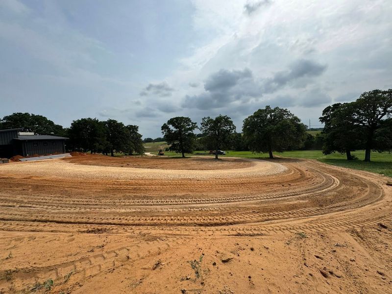 A curved dirt driveway under construction in a rural area with scattered trees under a cloudy sky.
