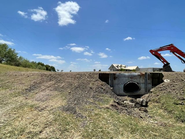 A concrete culvert pipe set into a gravel embankment under a clear blue sky, with a red excavator visible in the distance.