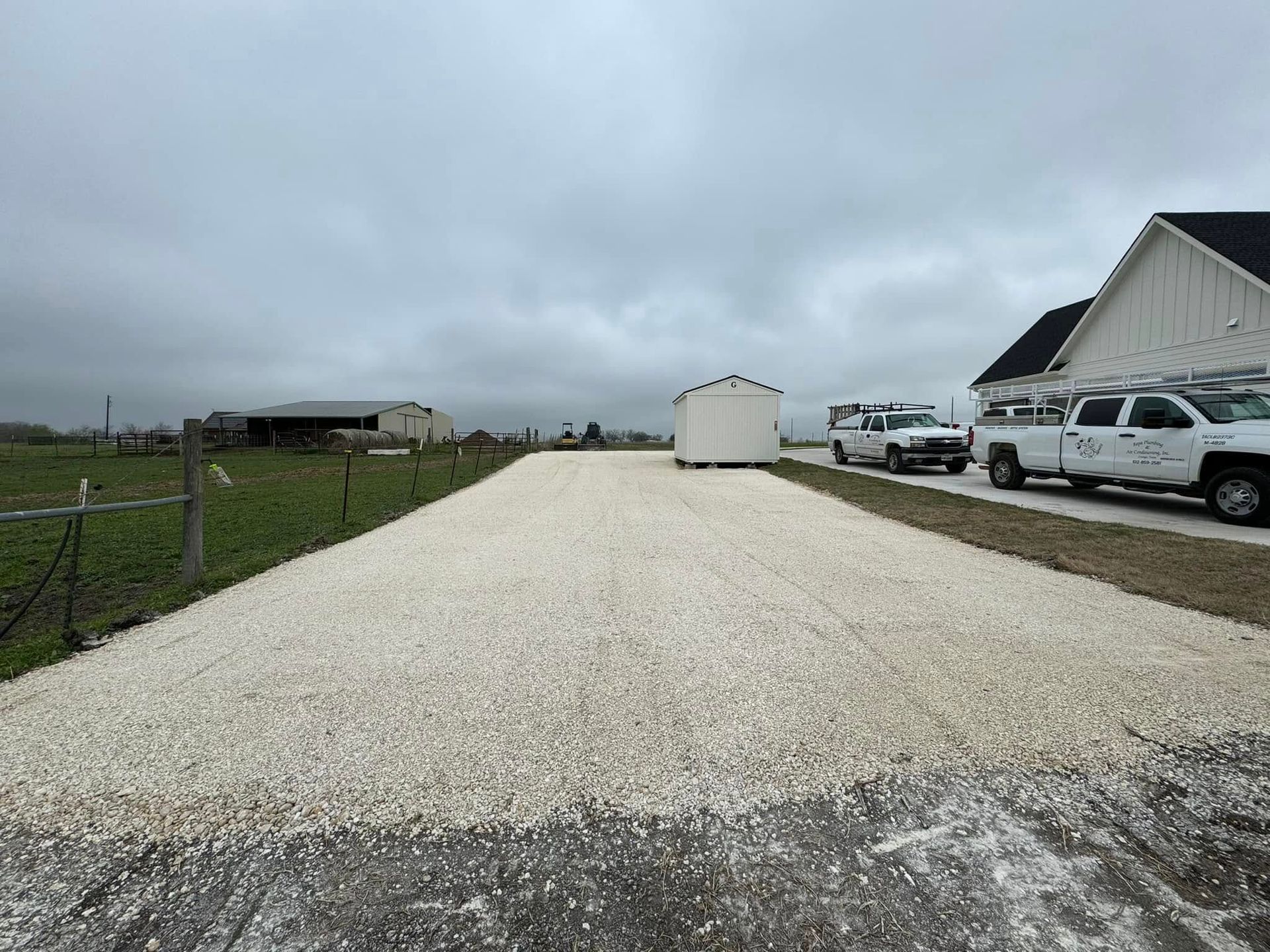 Gravel driveway leading to a white outbuilding and parked trucks under a cloudy sky.
