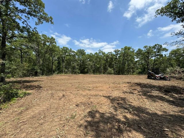 A cleared patch of dirt surrounded by green trees under a bright blue sky, with an ATV parked at the edge of the woods.