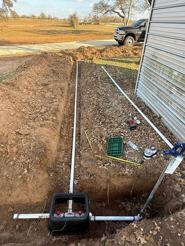 A valve box with PVC plumbing connected to irrigation lines in a dug trench next to a building.