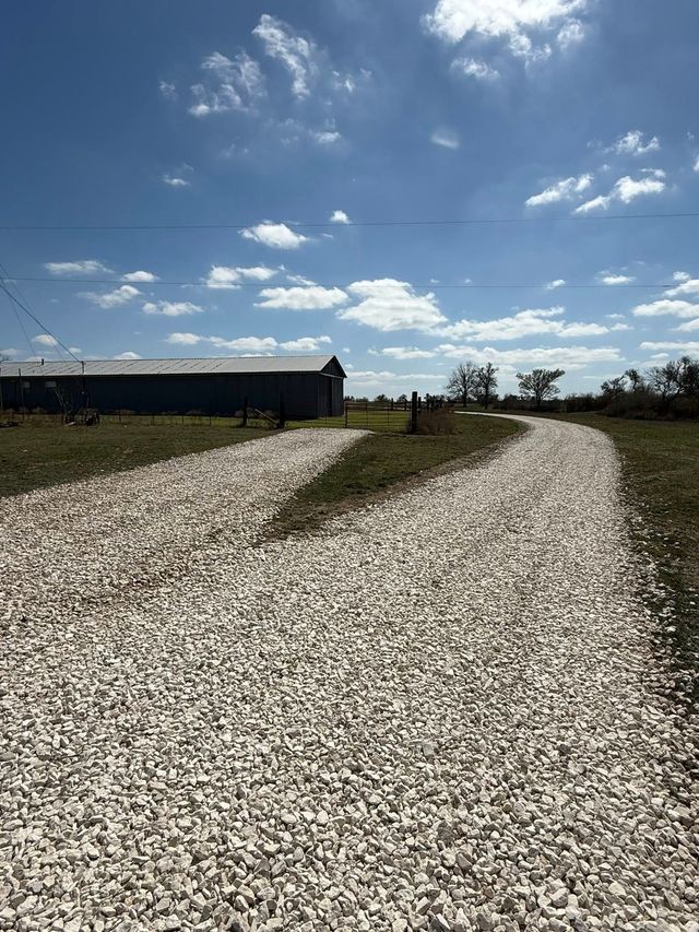 A white gravel driveway leads toward a long, dark storage building under a bright blue, partly cloudy sky.