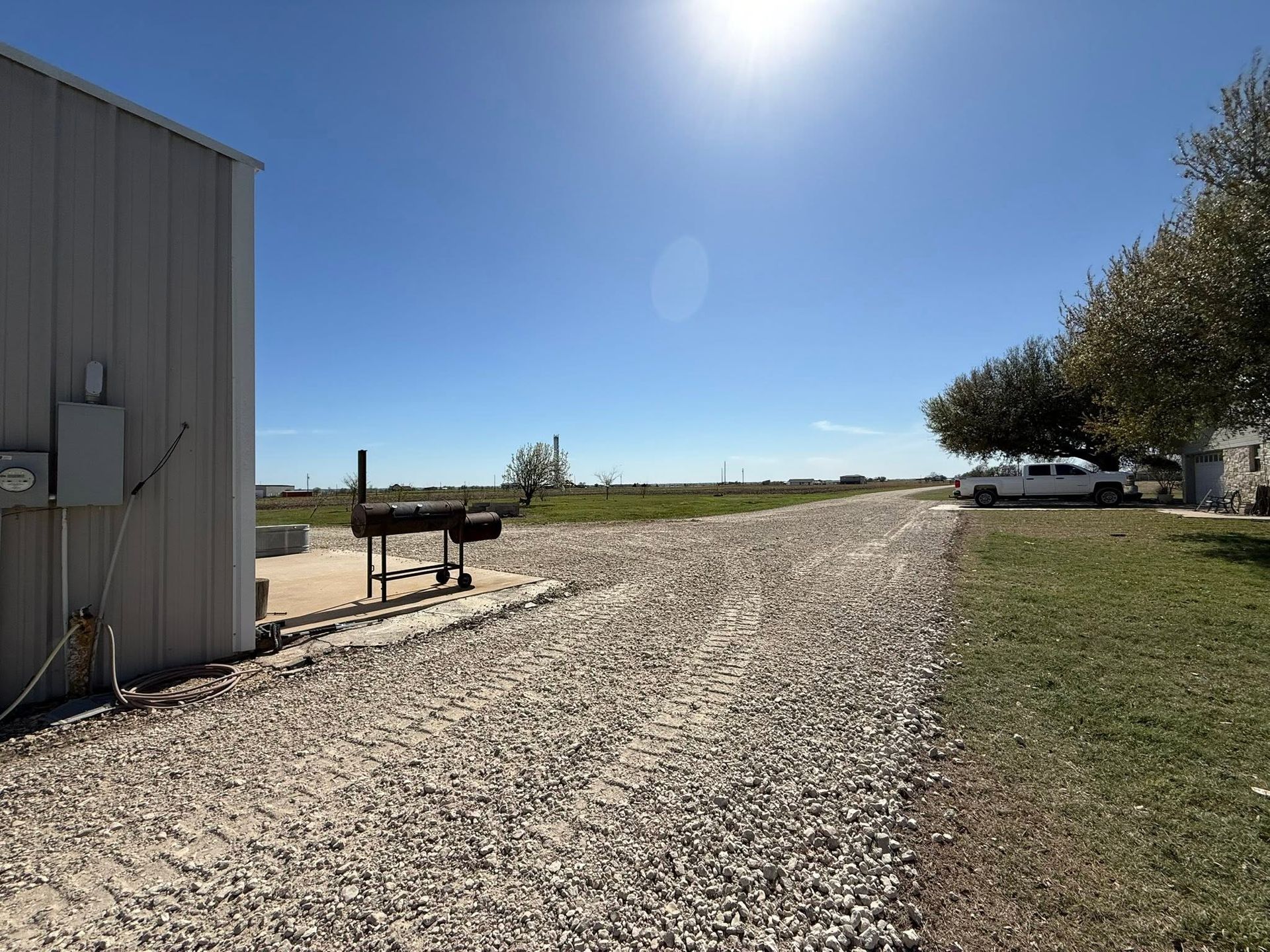 Gravel driveway beside a metal building under a bright blue sky, with open land in the distance.