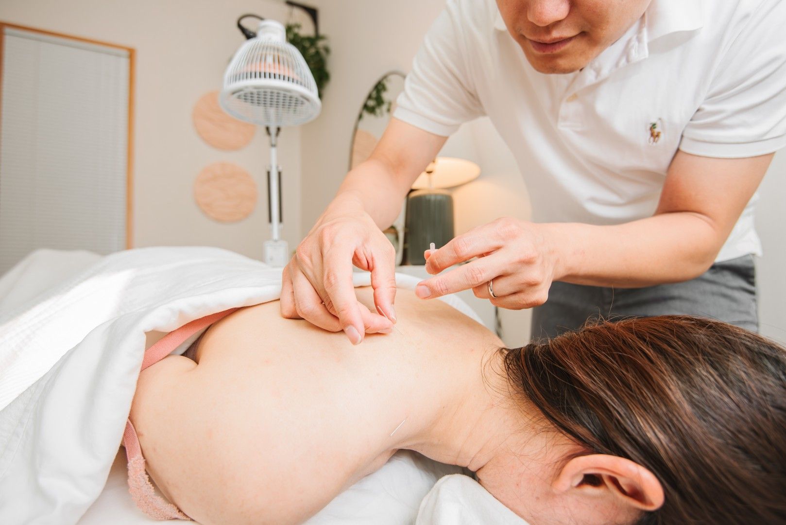 Acupuncturist inserting needles into patient's back. Light-filled room, white linens, and neutral walls.