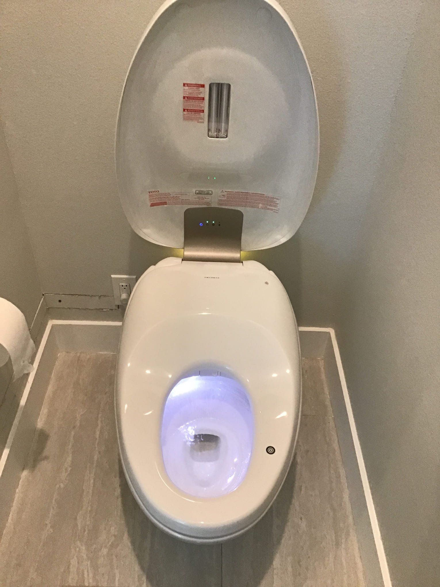 White, modern toilet with the lid up, glowing blue light inside the bowl, installed on a gray tiled floor.