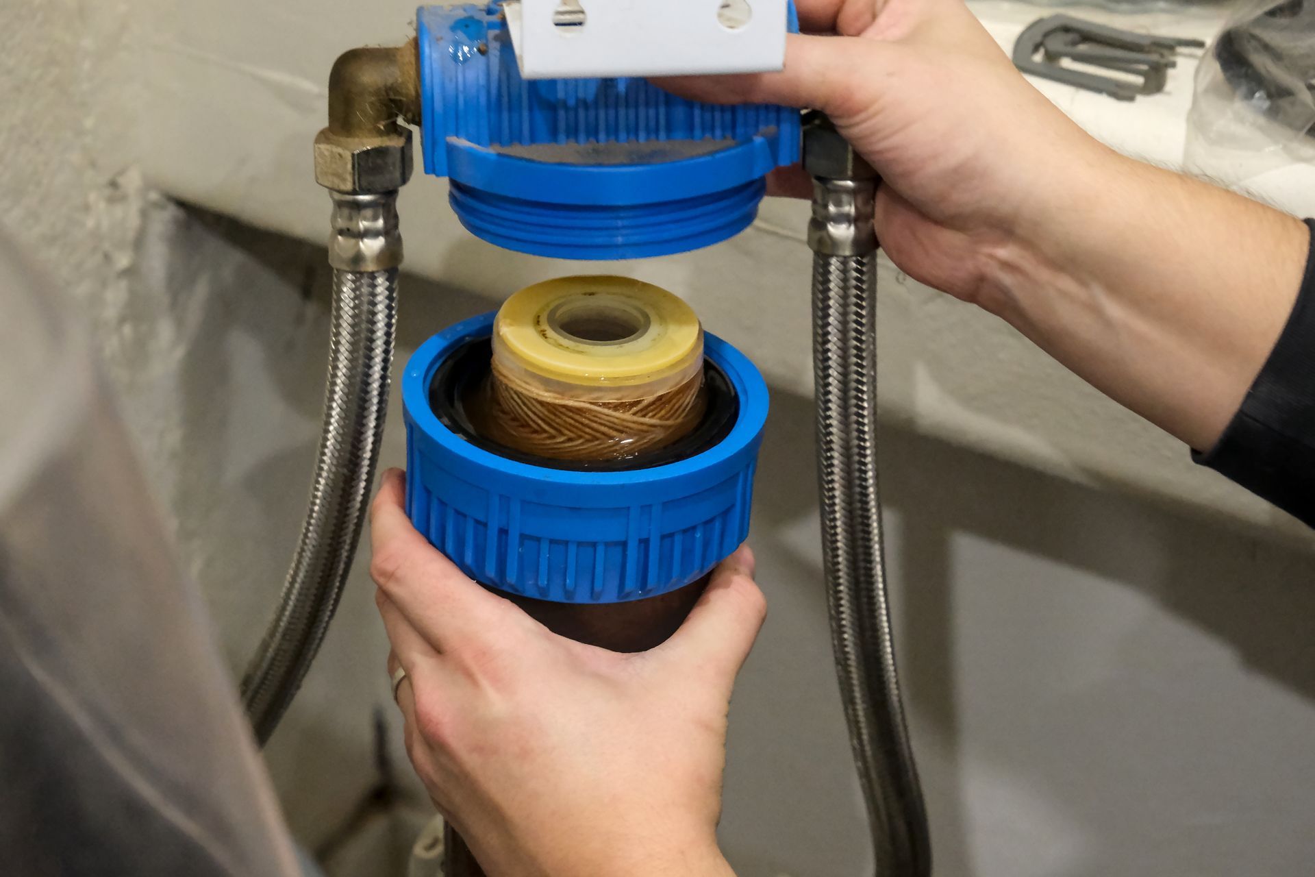 Hands removing a water filter cartridge from a blue housing connected to braided steel hoses