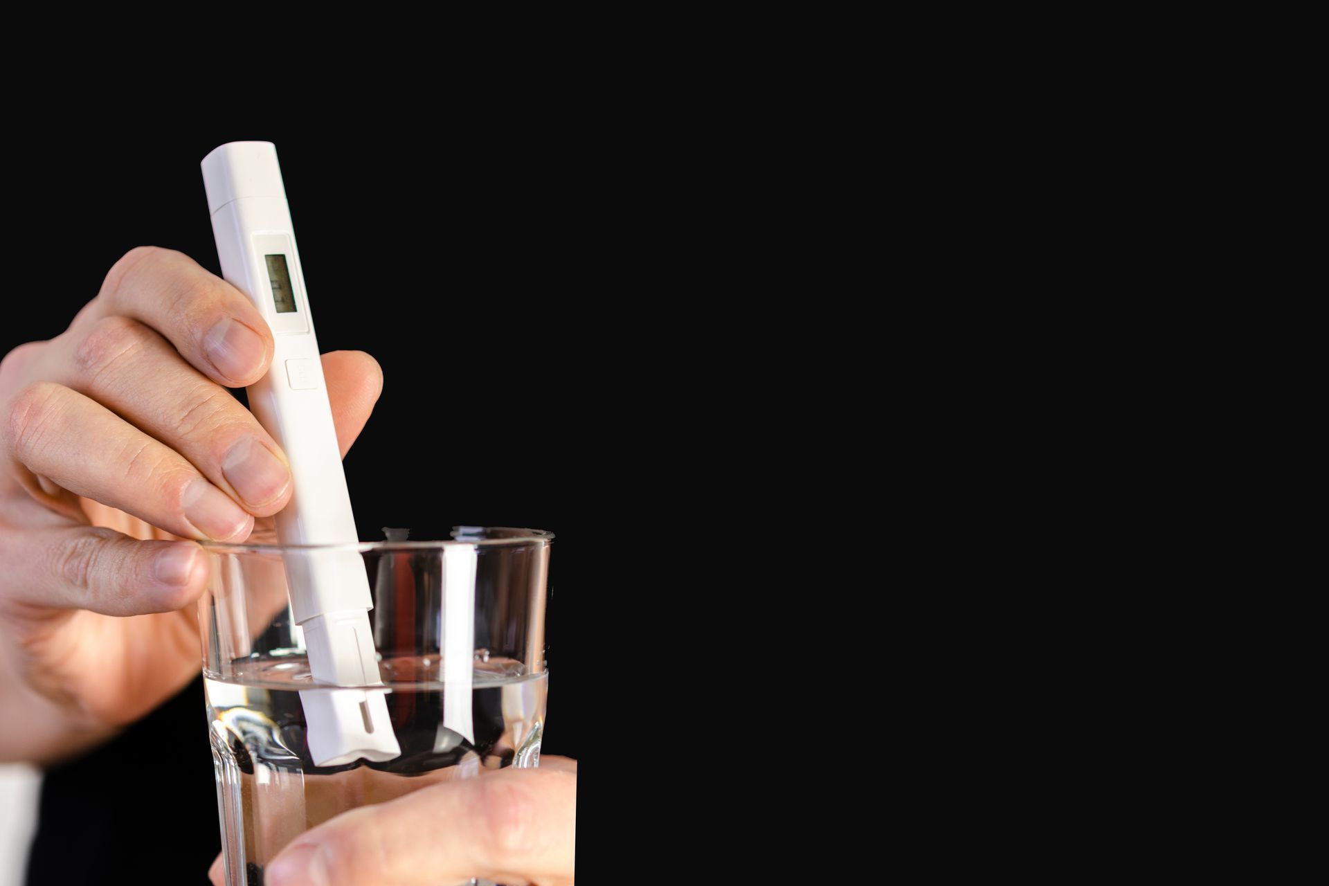 Person using a digital water quality tester in a glass of water against a black background. Person using a digital water quality tester in a glass of water against a black background.