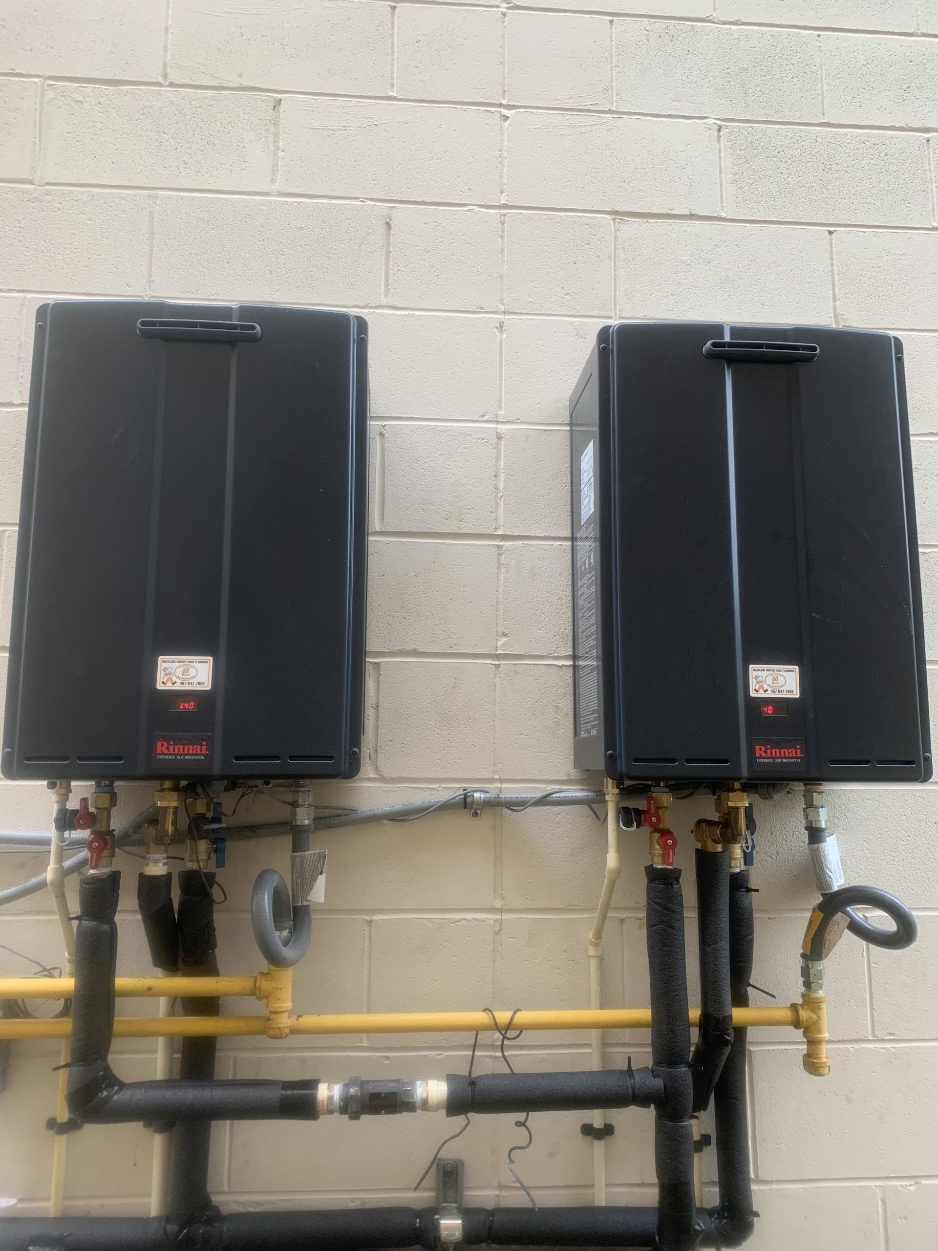 Two black tankless water heaters mounted on a concrete block wall, with yellow gas pipes and black water pipes.