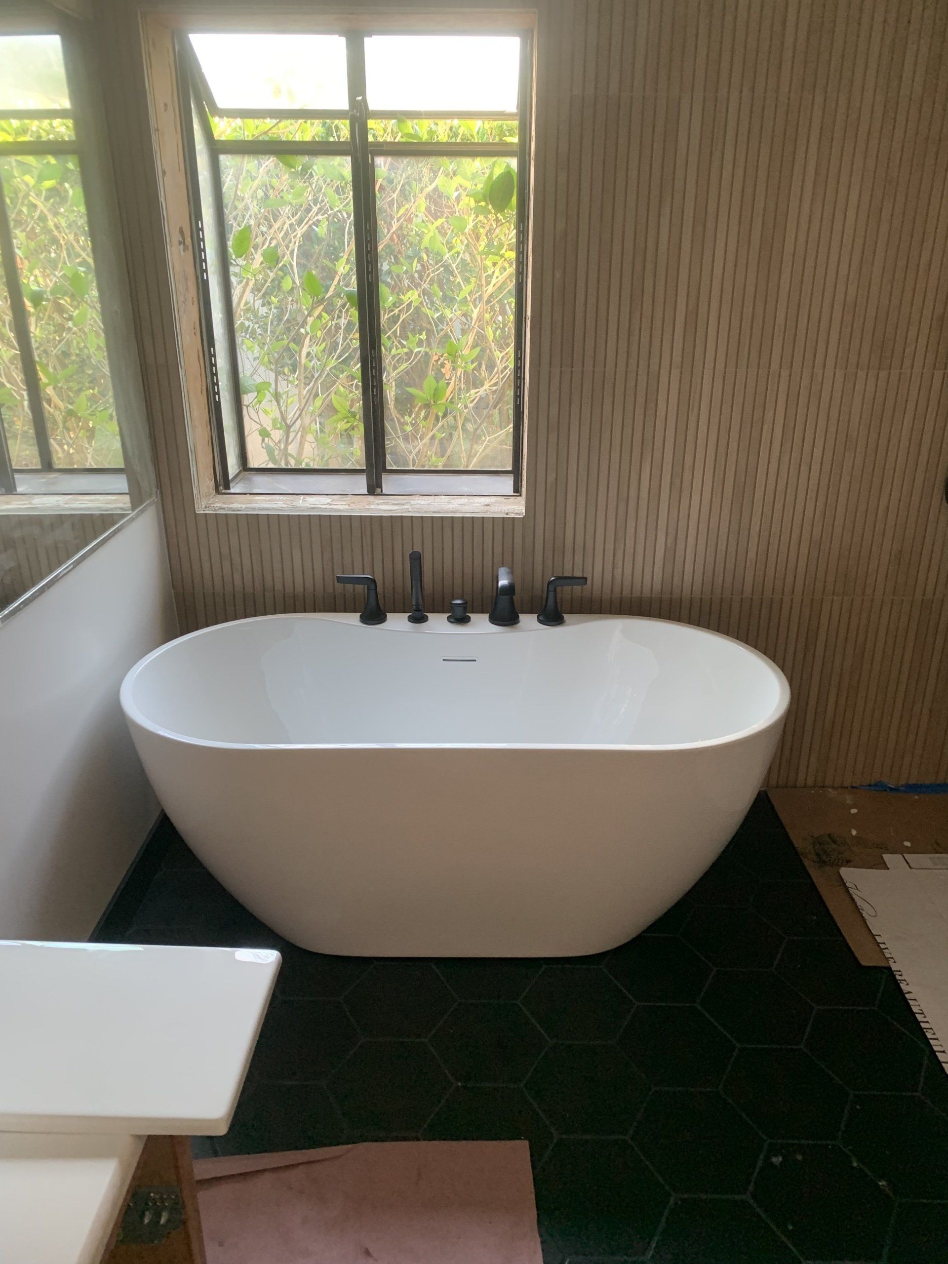 Freestanding white bathtub in a bathroom with dark green hexagon tile floor, and a window.