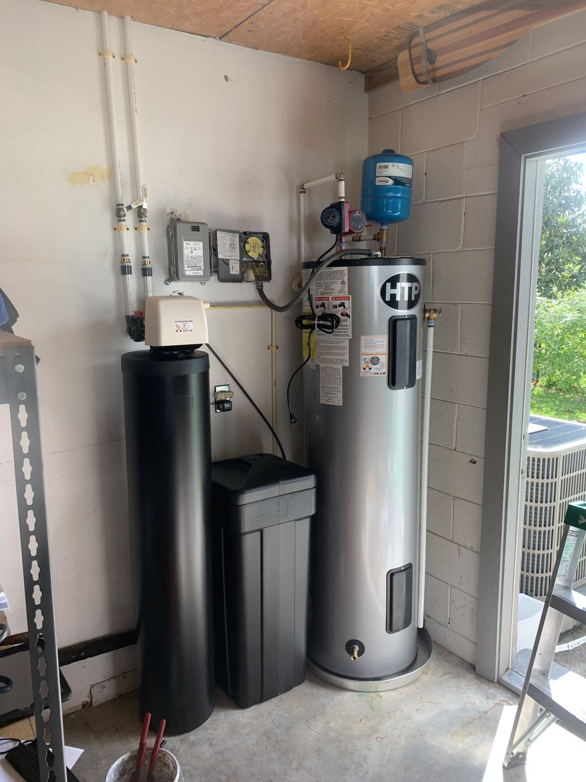 Water heater, water softener, and electrical boxes against a white cinder block wall in a garage.