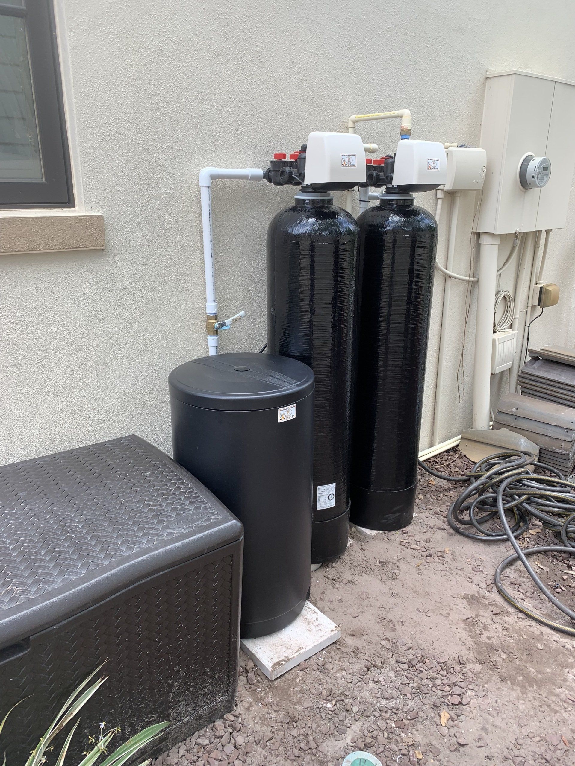 Outdoor water filtration system with two tall black tanks, a smaller black tank, and other equipment against a stucco wall.