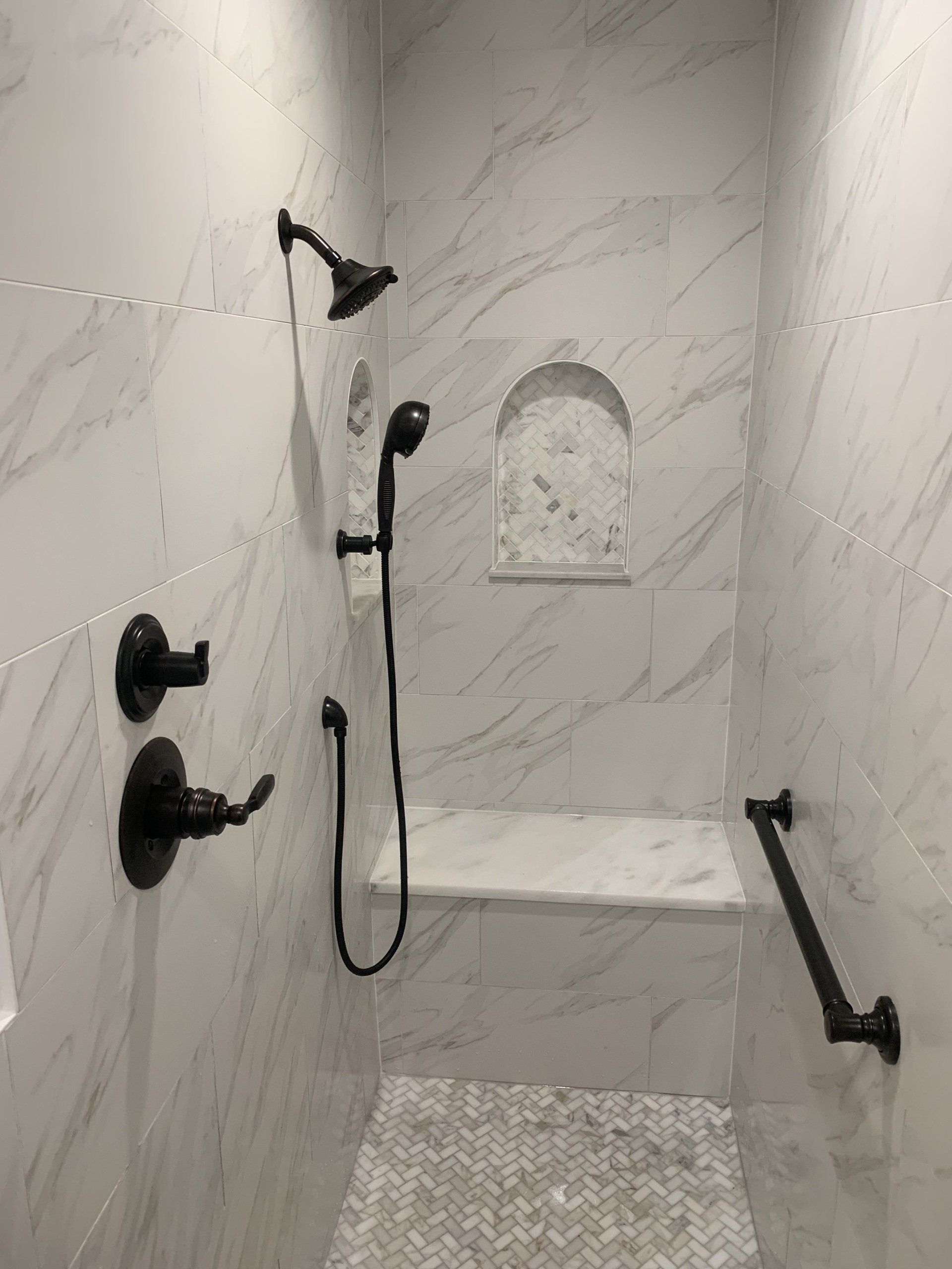 Marble-tiled shower with black fixtures; includes bench, hand-held shower, grab bar, and recessed shelf.