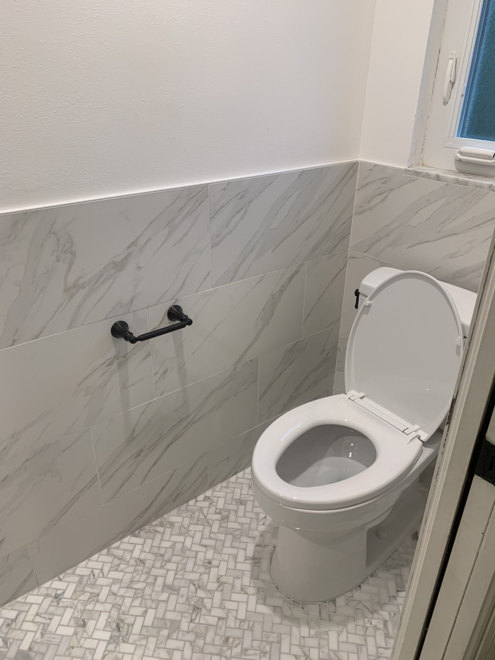 White toilet in a small bathroom with marble tile and mosaic floor. Black toilet paper holder.