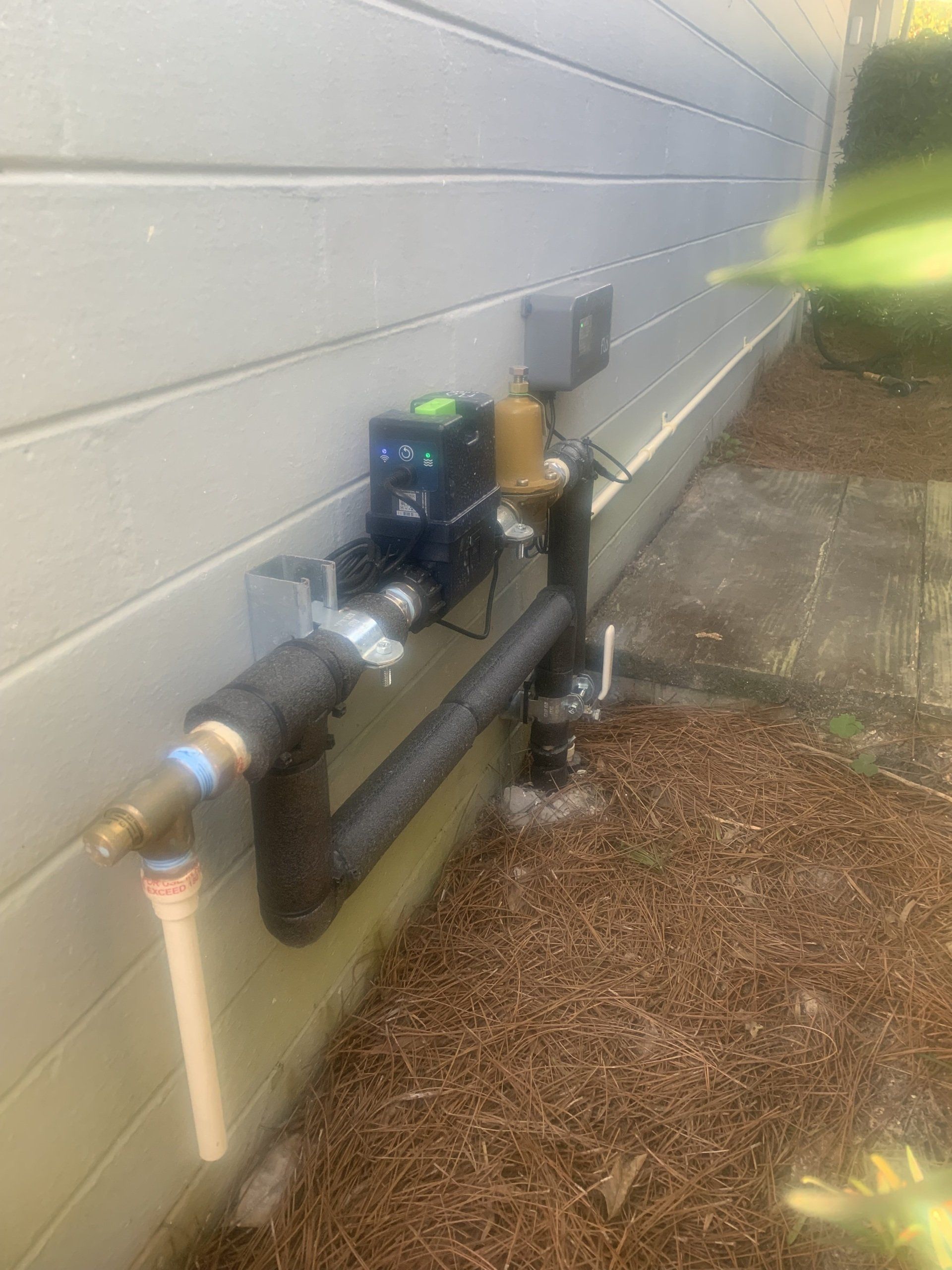 Black pipes and irrigation system components mounted on a light gray house exterior, set in mulch.