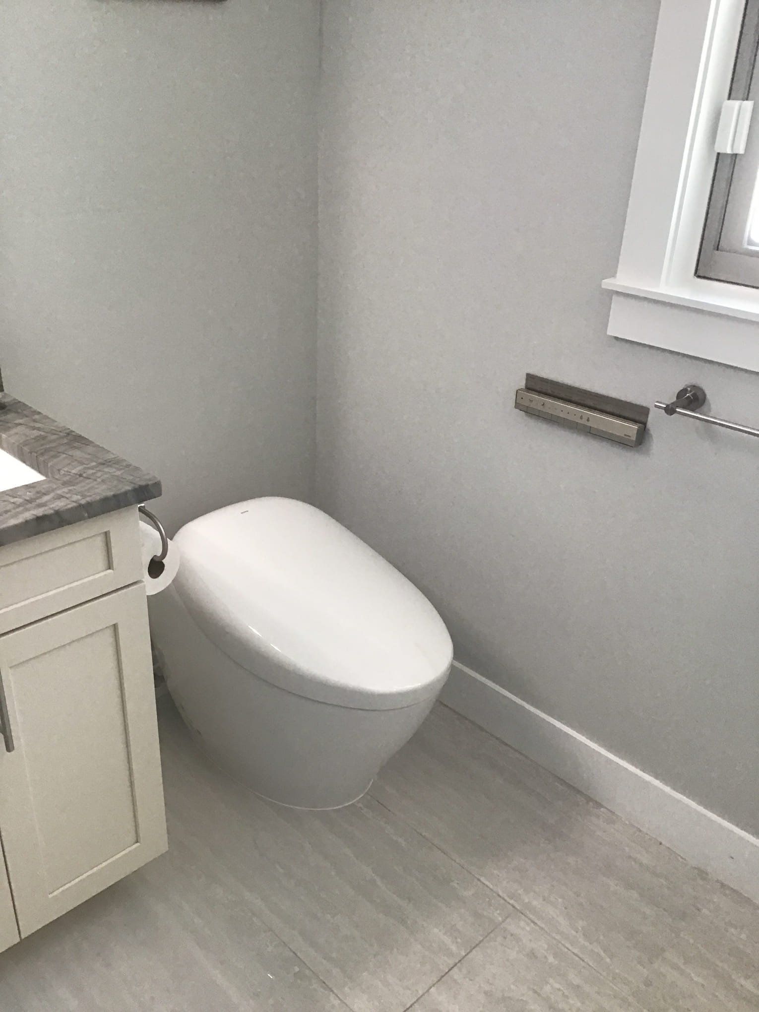 Modern white toilet in a gray-walled bathroom with grab bar and a countertop with cabinets.
