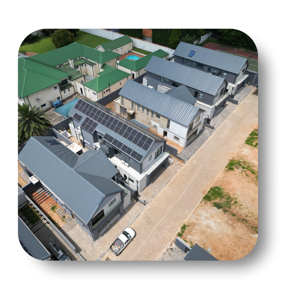 Aerial view of a modern residential complex with gray and green roofs and solar panels beside a dirt road.