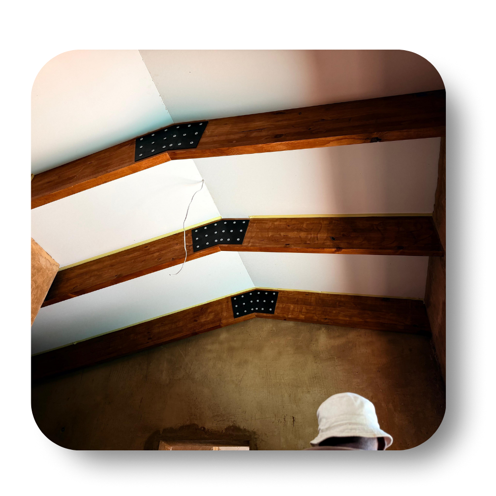 Wooden ceiling beams with black metal brackets against white walls, with a small white hard hat at bottom right