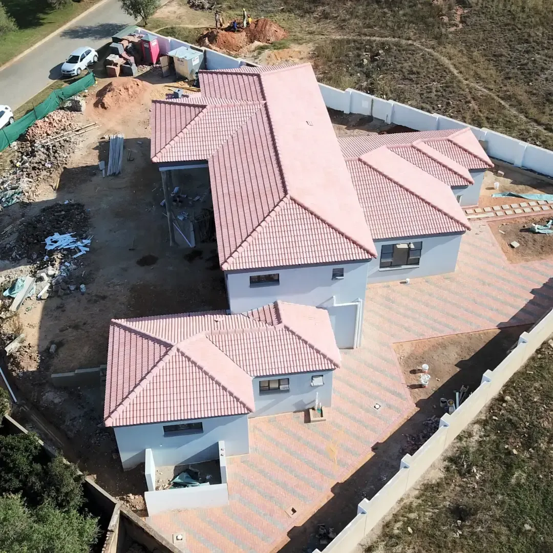 Aerial view of a white house with red tile roofs at a construction site, surrounded by dirt and fencing.