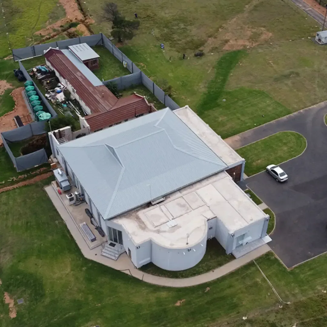 Aerial view of a white-roofed building with a curved front, paved driveway, and surrounding grassy grounds.