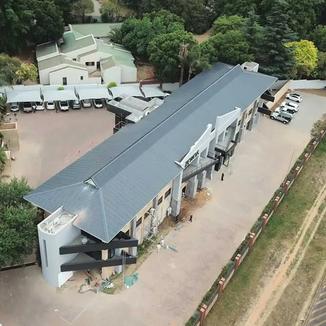 Aerial view of a long gray-roofed building with parked vehicles, trees, and an open courtyard.