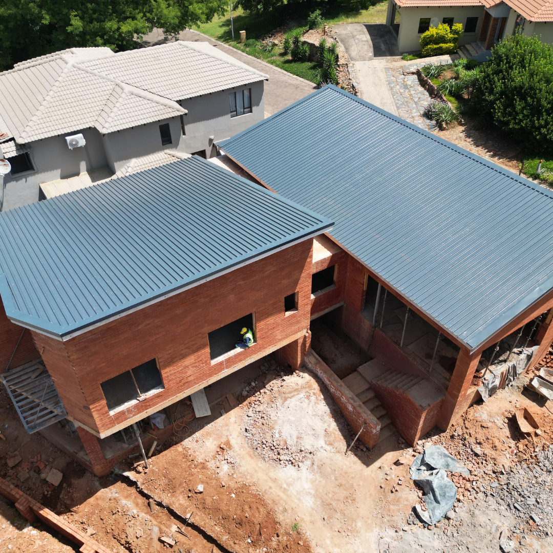 Aerial view of a brick house under construction with blue metal roofs and exposed dirt yard