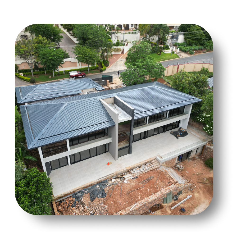 Aerial view of a modern gray-roofed building with a central walkway, surrounded by trees and construction dirt.