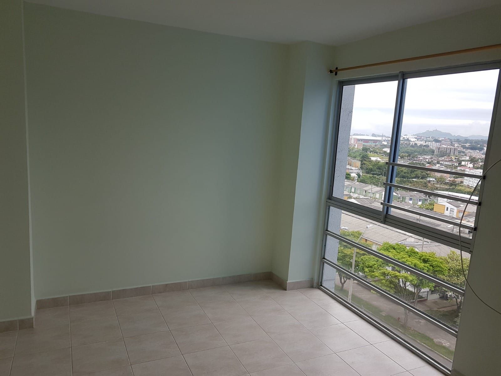 Habitación vacía con paredes de color verde pálido, suelo de baldosas y una gran ventana con vistas a la ciudad.