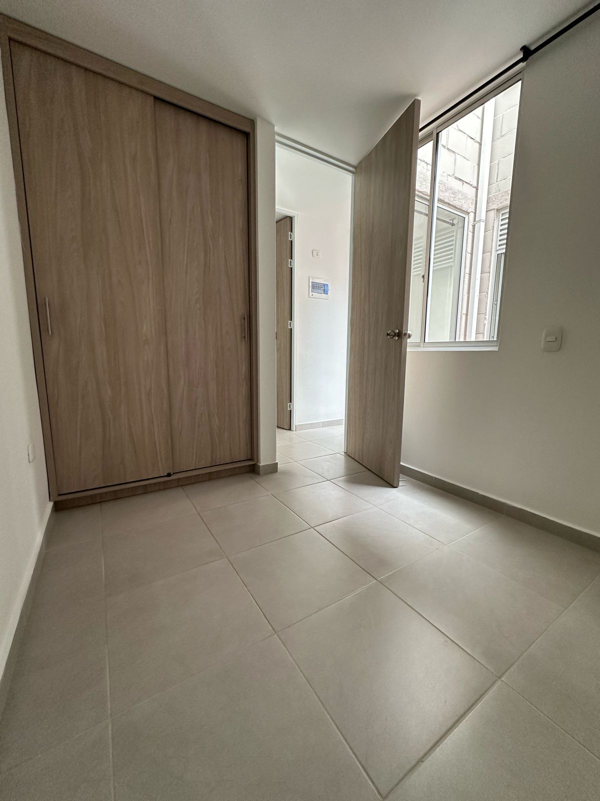 Habitación vacía con azulejos, puertas de armario de madera y una ventana que deja entrar la luz del día.