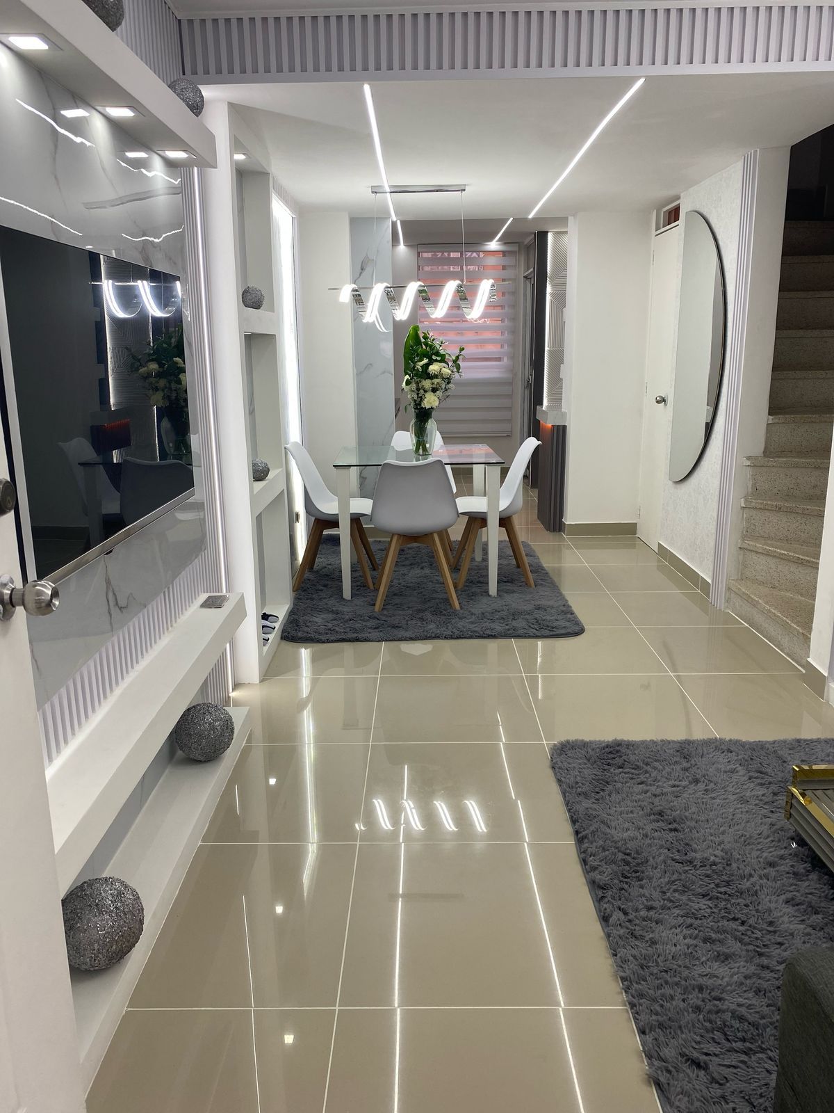Comedor moderno con mesa blanca, cuatro sillas, suelo de baldosas brillantes y alfombras grises en un pasillo luminoso.