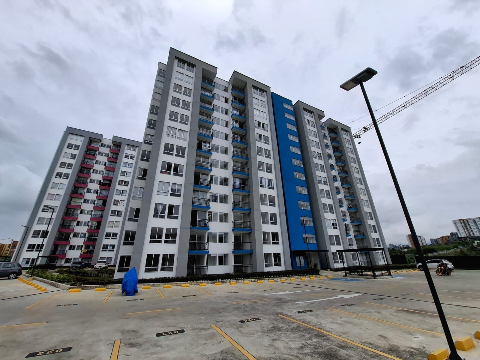 Edificios de apartamentos altos en un estacionamiento bajo un cielo nublado, con detalles azules y rojos en la fachada.