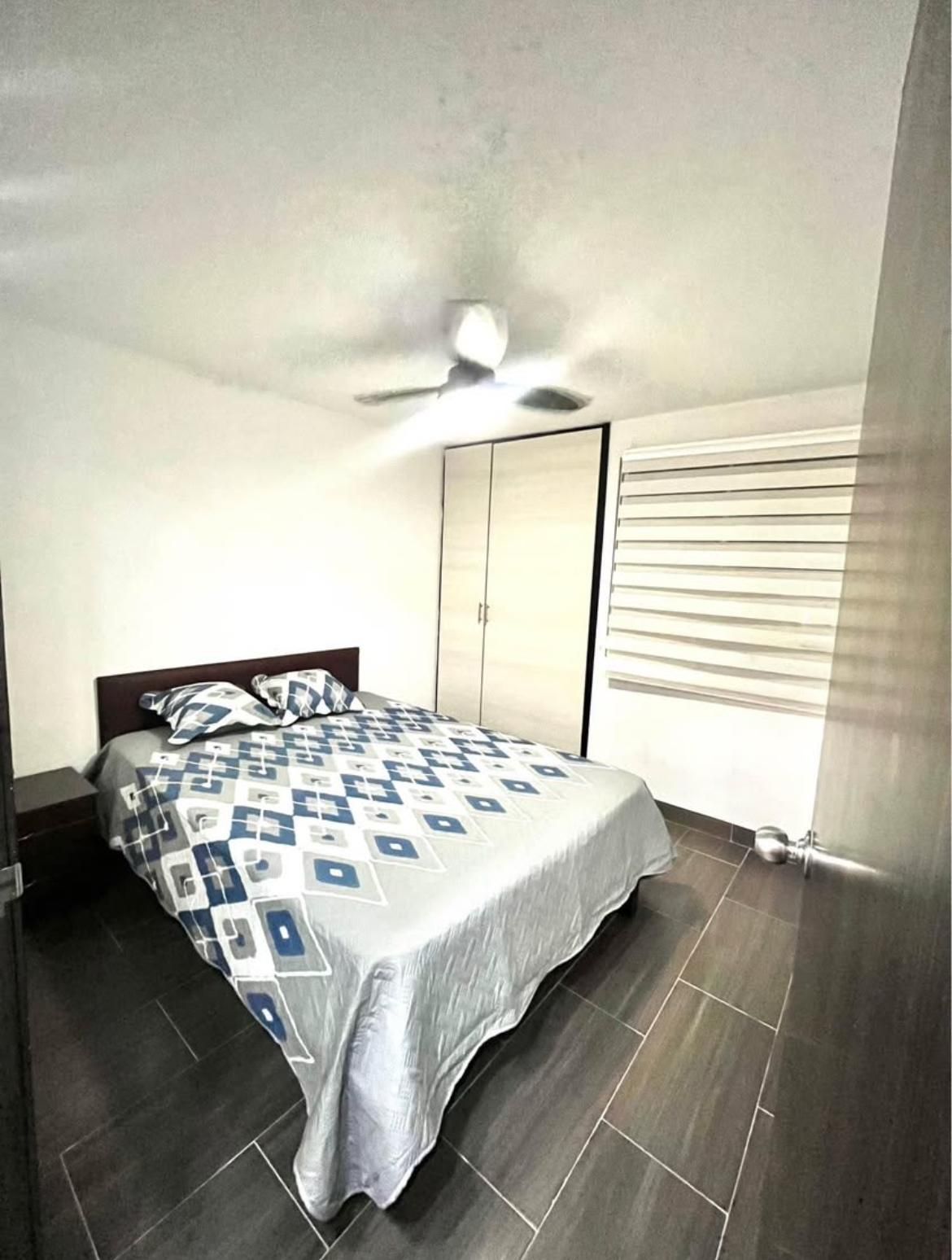 Habitación luminosa con cama, armario, ventilador de techo y persianas en las ventanas, sobre suelo de baldosas oscuras.