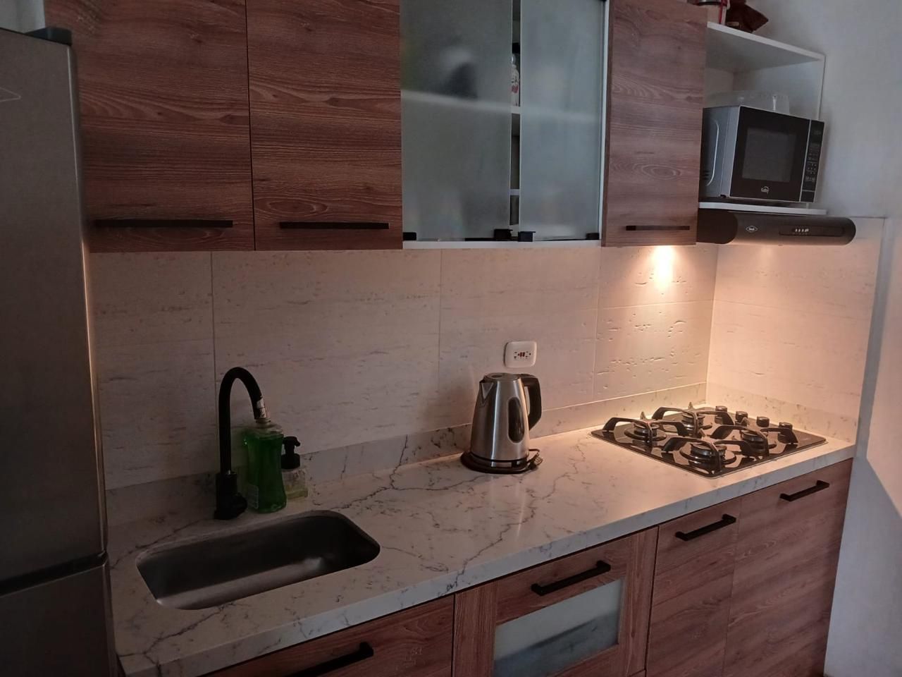 Cocina moderna con armarios de madera, fregadero, hervidor de agua y cocina de gas con iluminación bajo los armarios.