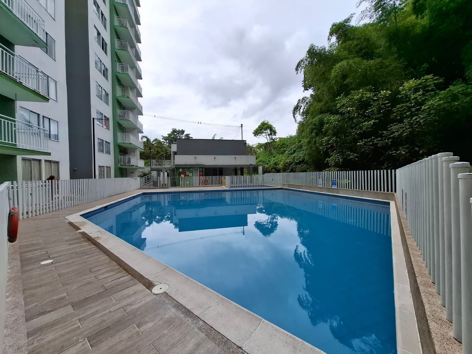 Piscina al aire libre junto a edificios de apartamentos, con agua azul y una terraza de madera bajo cielos nublados.
