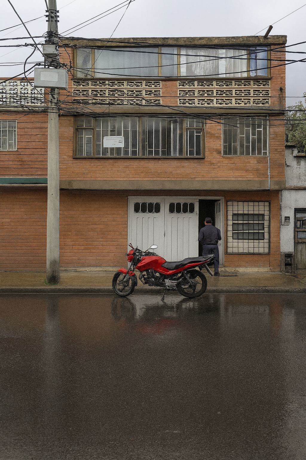 Calle lluviosa con un edificio de ladrillos y una motocicleta roja estacionada enfrente.