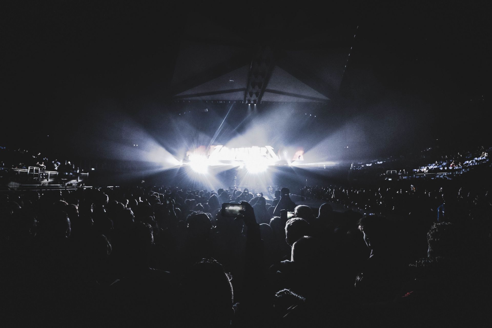 Crowd silhouetted at a night concert with bright white stage lights and city lights in the background