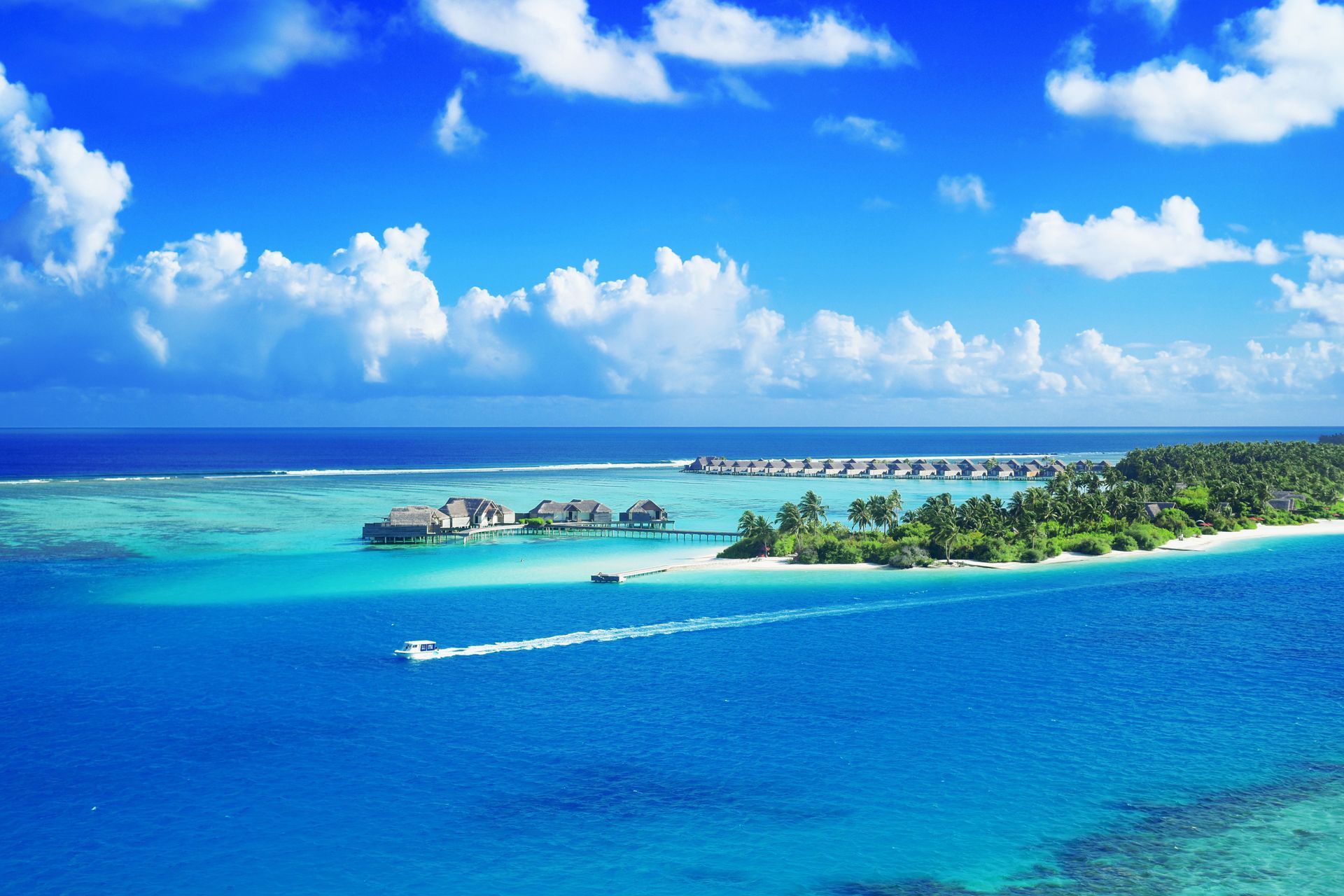 Tropical island resort with turquoise lagoon, white sand, palm trees, and a bright blue sky with clouds