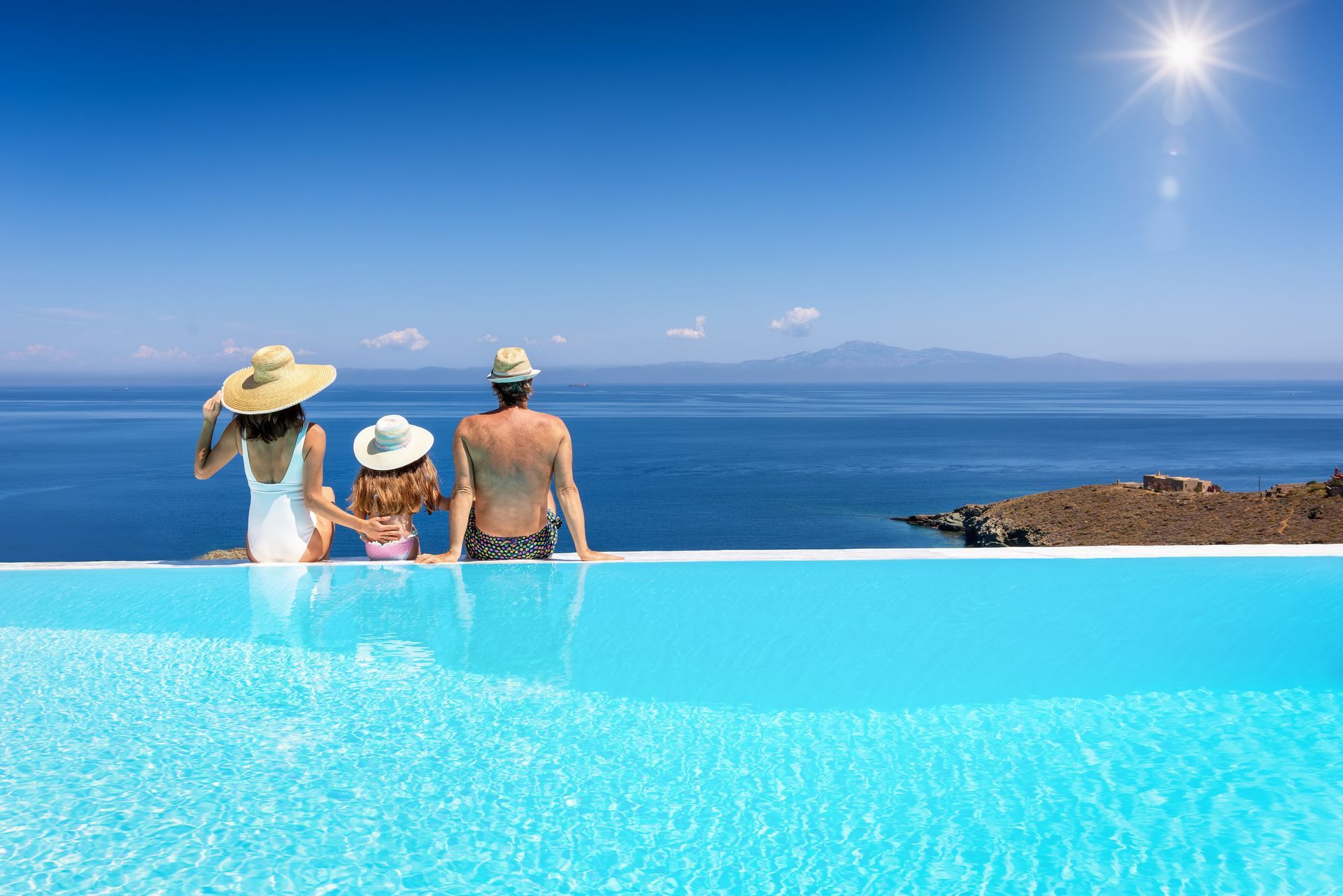 Three people sit at an infinity pool overlooking a bright blue ocean and clear sky.